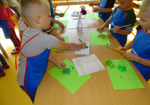 Dzieci stoją przy stoliku i malują liście zieloną farbą
